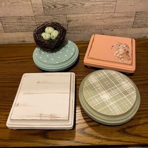 May include: Four different wooden decorative stands with various designs. One stand features a bird's nest with green eggs, another has a plaid pattern, a third has a whitewash finish, and the last one has a pink background with a bunny illustration.