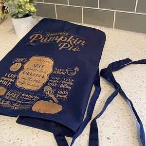 May include: A navy blue kitchen apron with gold text that reads "HOMEMADE Pumpkin Pie" and includes a recipe for pumpkin pie. The apron has two long ties.