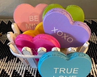 May include: A white picket fence basket filled with colorful wooden heart-shaped decorations. The hearts have various messages written on them, including "Be Mine", "XOXO", "Kiss", and "True Love".