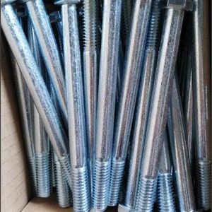 Puede incluir: Un primer plano de un conjunto de pernos de cabeza hexagonal plateados con vástagos roscados. Los pernos están cuidadosamente dispuestos dentro de una caja de cartón, mostrando su brillo metálico y diseño uniforme. Estos sujetadores son ideales para proyectos de construcción o reparación.