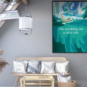 Puede incluir: Un cuadro enmarcado con un surfista en olas turquesas y azules con el texto "I'm working on a new me". La obra de arte cuelga sobre un banco de mimbre con cojines. También se ven una lámpara decorativa y hierba de la pampa.