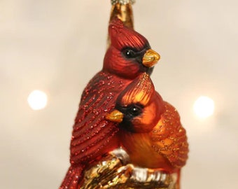 May include: A glass Christmas ornament featuring two red cardinals perched on a gold branch. The cardinals are detailed with red, orange, and white paint.