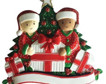 May include: A Christmas bauble depicting two people in red and white pyjamas holding presents in front of a green Christmas tree with red baubles. The bauble has a white banner at the bottom for personalisation.