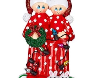May include: A Christmas bauble depicting a couple in red and white pyjamas with white polka dots, holding a string of Christmas lights and a wreath. The couple are wearing Santa hats and have blue eyes. The bauble is set against a white background.