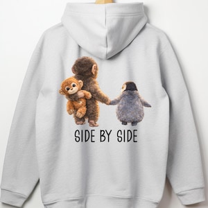 May include: A light gray hoodie featuring a whimsical illustration of three animal characters. A brown monkey holds a smaller monkey, and they are holding hands with a gray penguin. The text "SIDE BY SIDE" is printed below the characters.