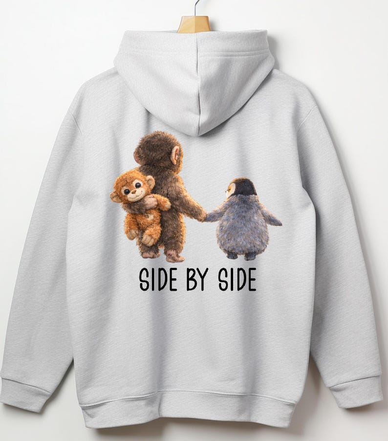 May include: A light gray hoodie featuring a whimsical illustration of three animal characters. A brown monkey holds a smaller monkey, and they are holding hands with a gray penguin. The text "SIDE BY SIDE" is printed below the characters.