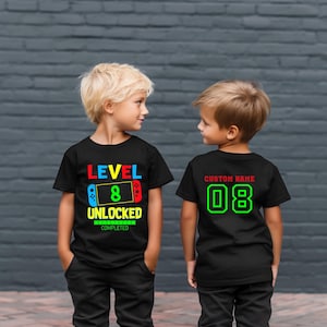May include: Two black t-shirts for kids. The front of one shirt says "Level 8 Unlocked Completed" with a colorful video game controller graphic. The back of the other shirt says "Custom Name 08" in green.
