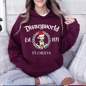 Pu&ograve; includere: Una felpa con cappuccio Disneyworld bordeaux con una grafica frontale. La grafica presenta il testo "Disneyworld Est. 1971 FLORIDA" con un personaggio dei cartoni animati con un cappello da Babbo Natale. La felpa ha un cappuccio con coulisse e una tasca frontale.