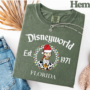 May include: An olive green t-shirt with the text "Disneyworld Est. 1971 Florida". The shirt features a cartoon character wearing a Santa hat and surrounded by a wreath of lights. A silver necklace is draped across the shirt.