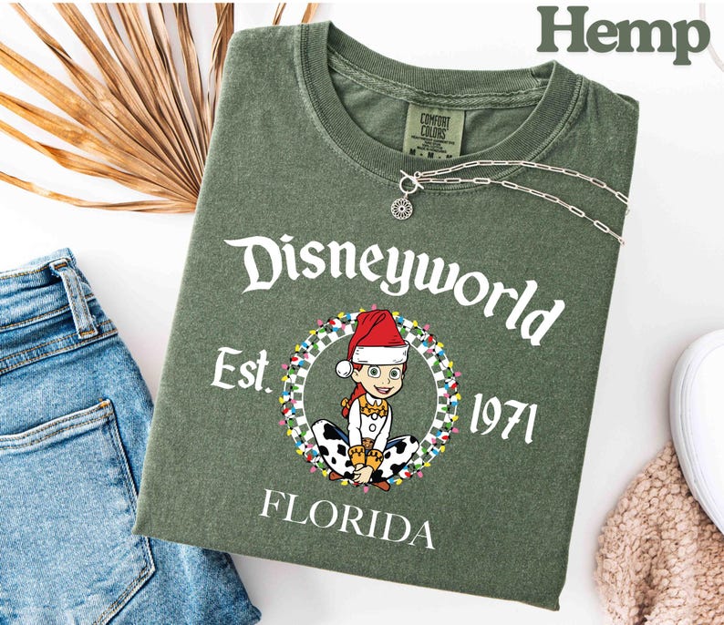 May include: An olive green t-shirt with the text "Disneyworld Est. 1971 Florida". The shirt features a cartoon character wearing a Santa hat and surrounded by a wreath of lights. A silver necklace is draped across the shirt.