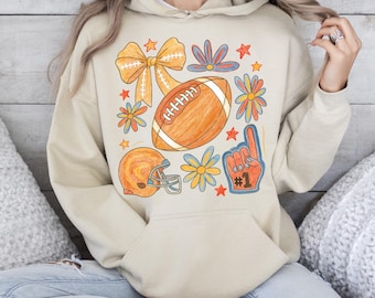 May include: A beige hoodie featuring a football-themed graphic. The design includes an orange football, a helmet, a bow, flowers, stars, and a foam finger with the number one. The hoodie has a front pocket and a drawstring hood.