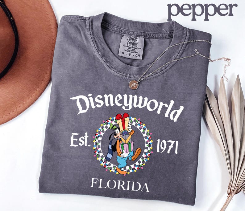 May include: Charcoal grey t-shirt with "Disneyworld" in white script, featuring a cartoon Goofy character holding gifts. The shirt includes "Est. 1971" and "Florida" in white text. A silver necklace with a pendant is on the shirt.