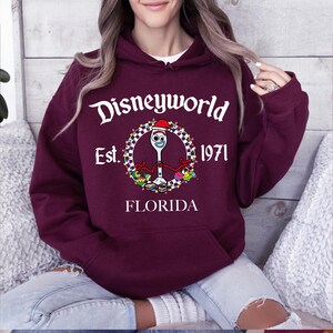 Pu&ograve; includere: Una felpa con cappuccio Disneyworld bordeaux con una grafica frontale. La grafica presenta il testo "Disneyworld Est. 1971 Florida" attorno a un personaggio dei cartoni animati. Il personaggio &egrave; una forchetta con un volto e un cappello da Babbo Natale.
