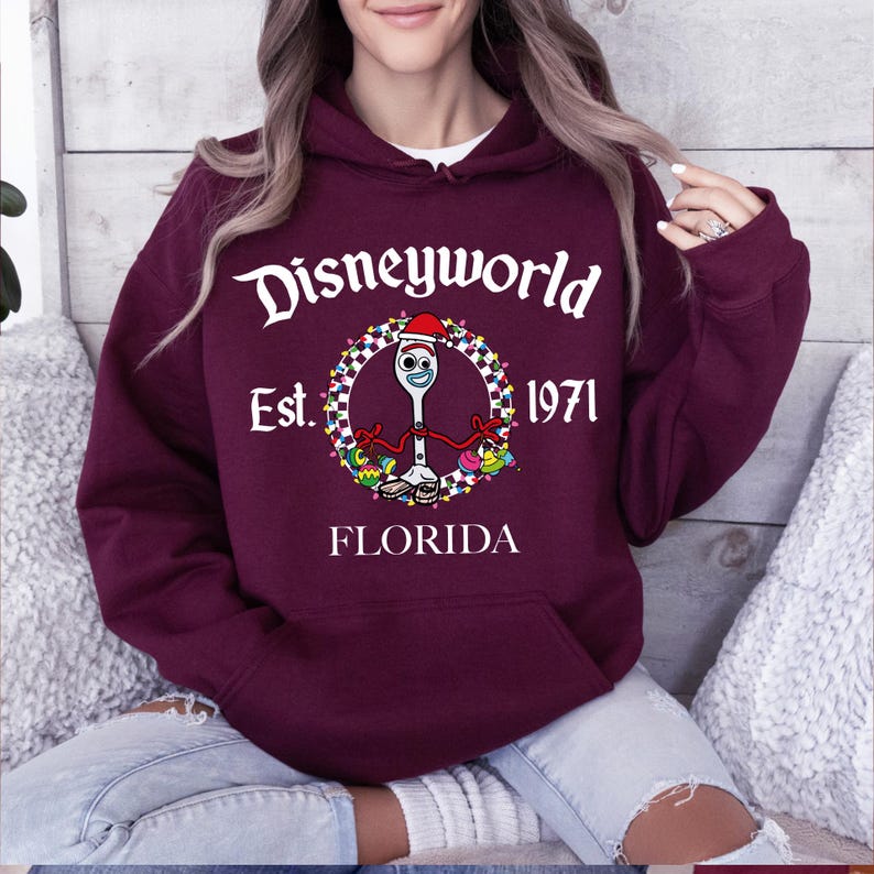 Pu&ograve; includere: Una felpa con cappuccio Disneyworld bordeaux con una grafica frontale. La grafica presenta il testo "Disneyworld Est. 1971 Florida" attorno a un personaggio dei cartoni animati. Il personaggio &egrave; una forchetta con un volto e un cappello da Babbo Natale.