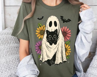 May include: Olive green t-shirt featuring a Halloween-themed graphic. The design includes a black cat peeking out from under a ghost sheet, surrounded by colorful flowers and bats. A fun and festive design for the spooky season.