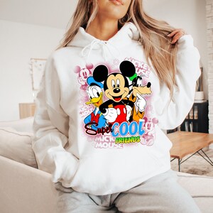 Puede incluir: Sudadera con capucha blanca con un gr&aacute;fico colorido de Mickey Mouse, el Pato Donald y Goofy. El gr&aacute;fico incluye el texto "Super Cool Mickey Mouse Friends". El dise&ntilde;o est&aacute; sobre un fondo rosa con texto de Mickey Mouse.
