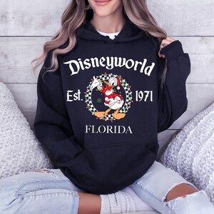 Pu&ograve; includere: Una felpa con cappuccio blu navy con la scritta "Disneyworld Est. 1971 FLORIDA". Il design presenta un personaggio dei cartoni animati di Paperino in abito natalizio, circondato da una ghirlanda di luci colorate e un bordo a scacchi.