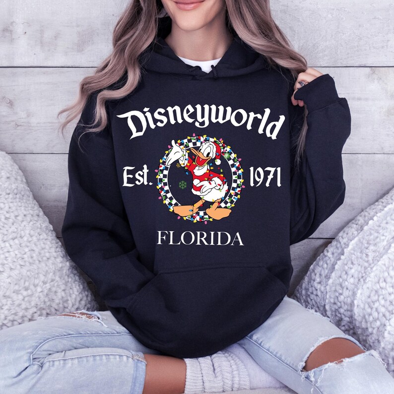 Pu&ograve; includere: Una felpa con cappuccio blu navy con la scritta "Disneyworld Est. 1971 FLORIDA". Il design presenta un personaggio dei cartoni animati di Paperino in abito natalizio, circondato da una ghirlanda di luci colorate e un bordo a scacchi.
