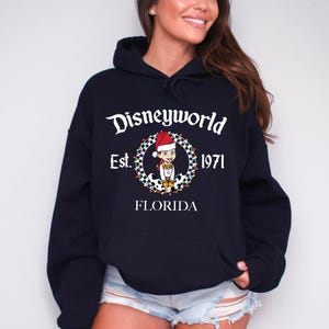 Pu&ograve; includere: Una felpa con cappuccio blu navy con una tasca frontale. La felpa presenta una grafica di un personaggio dei cartoni animati con un cappello da Babbo Natale, circondato da una ghirlanda, con la scritta "Disneyworld Est. 1971 FLORIDA".