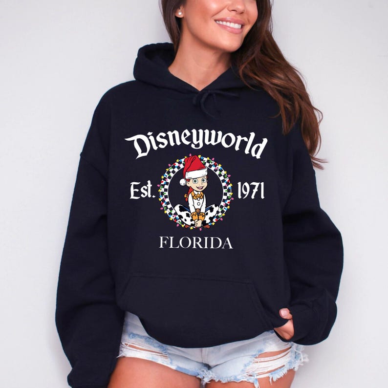 Pu&ograve; includere: Una felpa con cappuccio blu navy con una tasca frontale. La felpa presenta una grafica di un personaggio dei cartoni animati con un cappello da Babbo Natale, circondato da una ghirlanda, con la scritta "Disneyworld Est. 1971 FLORIDA".