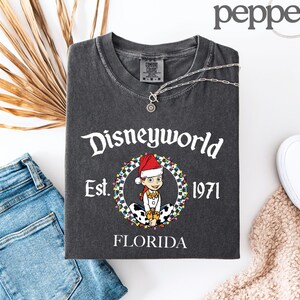 May include: Dark grey t-shirt with a Disneyworld graphic. The graphic features a cartoon character wearing a Santa hat, surrounded by a wreath of lights, with the text "Disneyworld Est. 1971 Florida". A silver necklace is draped over the shirt.