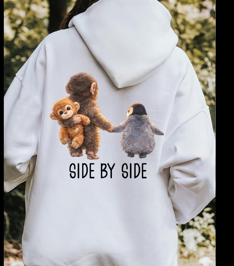 May include: White hoodie featuring a whimsical illustration of three animal figures: a brown monkey holding a smaller monkey, and a gray penguin-like creature. The phrase "SIDE BY SIDE" is printed below the figures in black. The hoodie has a hood and long sleeves.