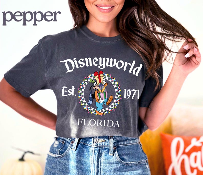 May include: Dark grey t-shirt with a Disney World graphic featuring Goofy holding gifts, surrounded by a circular design with the text "Disneyworld Est. 1971 Florida". The shirt is paired with blue denim jeans.