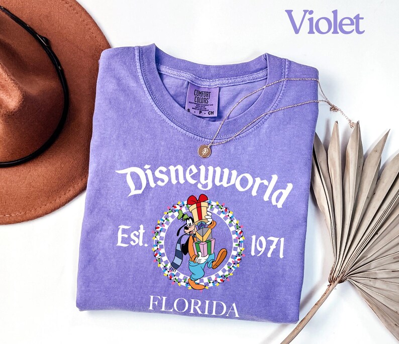 May include: A violet t-shirt with a Disneyworld graphic featuring Goofy holding gifts. The shirt includes the text "Disneyworld Est. 1971 Florida". A gold necklace and a brown hat are also in the image.
