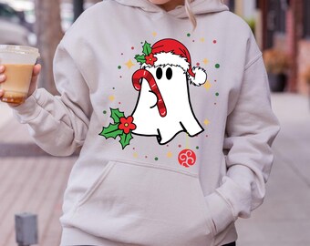 May include: A beige hoodie featuring a Christmas ghost graphic. The ghost wears a Santa hat and holds a candy cane. Holly and decorative dots surround the ghost. The hoodie has a front pocket and a drawstring hood.