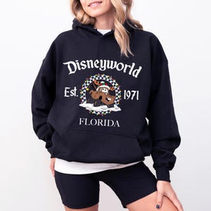 Pu&ograve; includere: Felpa con cappuccio nera con la scritta "Disneyworld" e un disegno di un'auto dei cartoni animati con un cappello di Babbo Natale. La felpa ha anche la scritta "Est. 1971" e "Florida" stampata.