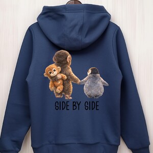 May include: Navy blue hoodie featuring a whimsical illustration of a monkey holding a smaller monkey, and a penguin holding hands. The phrase "SIDE BY SIDE" is printed below the image in black letters. The hoodie has a hood and a drawstring.