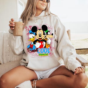 Puede incluir: Una sudadera con capucha color crema con un gr&aacute;fico de Mickey Mouse, Donald Duck y Goofy con el texto "Super Cool Mickey Friends". El dise&ntilde;o incluye acentos rosas y las palabras "Mickey Mouse" en un patr&oacute;n circular. La sudadera tiene una capucha con cord&oacute;n.