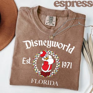 May include: Espresso-coloured t-shirt with a Donald Duck graphic in a Santa hat, and the text "Disneyworld Est. 1971 Florida." The shirt has a crew neck and short sleeves. A silver necklace is draped across the shirt.
