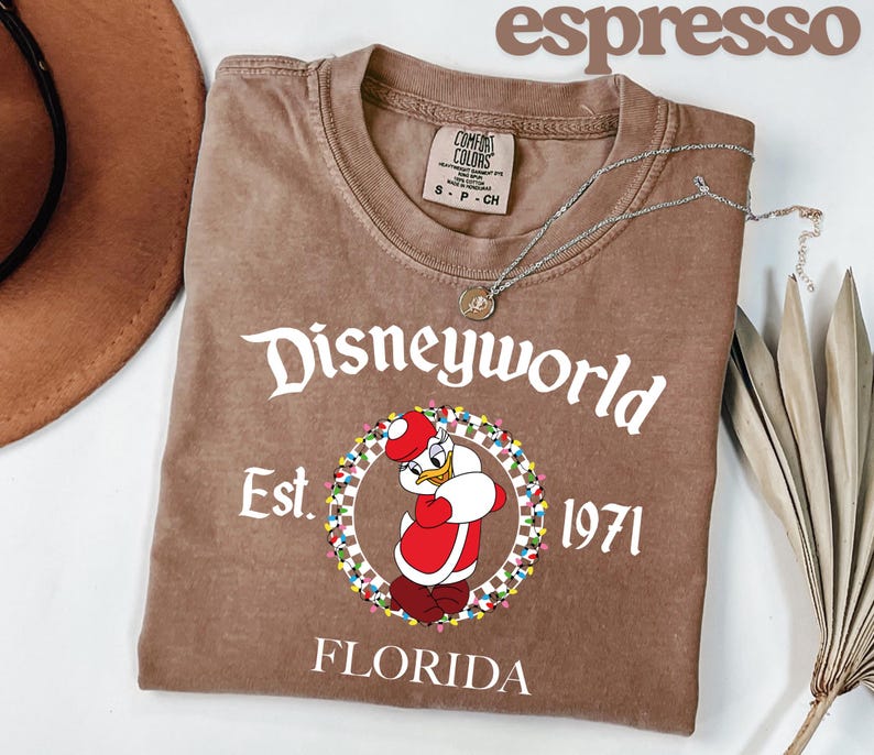 May include: Espresso-coloured t-shirt with a Donald Duck graphic in a Santa hat, and the text "Disneyworld Est. 1971 Florida." The shirt has a crew neck and short sleeves. A silver necklace is draped across the shirt.