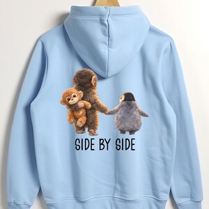 May include: Light blue hoodie featuring a graphic of three cartoon animals: a monkey holding a smaller monkey, and a penguin. The text "SIDE BY SIDE" is printed below the animals in black. The hoodie is hanging on a wooden hanger.