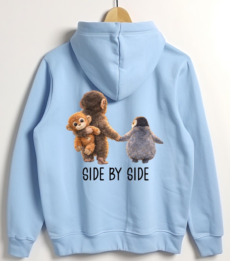 May include: Light blue hoodie featuring a graphic of three cartoon animals: a monkey holding a smaller monkey, and a penguin. The text "SIDE BY SIDE" is printed below the animals in black. The hoodie is hanging on a wooden hanger.