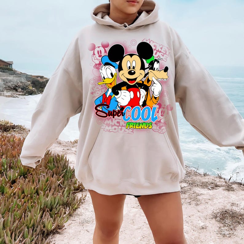 Puede incluir: Sudadera con capucha beige con un gr&aacute;fico de Mickey Mouse, el Pato Donald y Goofy. El texto "Super Cool Friends" est&aacute; debajo de los personajes. El dise&ntilde;o incluye siluetas rosas de Mickey Mouse y las palabras "Mickey Mouse".