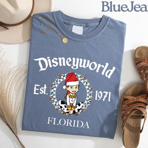 May include: A blue t-shirt with the text "Disneyworld Est. 1971 Florida" and a cartoon character wearing a Santa hat. The shirt is paired with brown sandals and a beige hat.