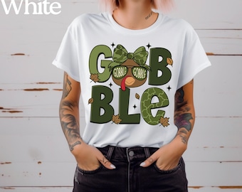 May include: White t-shirt with a Thanksgiving-themed graphic. The design features the word "GOBBLE" in large, green letters, with a cartoon turkey wearing sunglasses and a bow tie. The word "White" is printed at the top.
