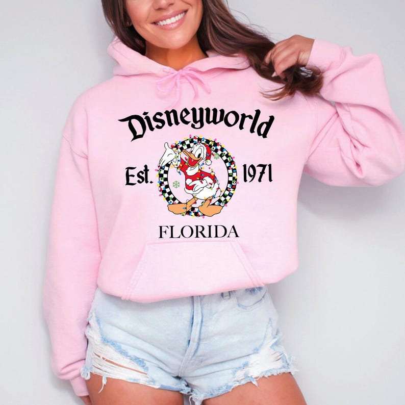 Pu&ograve; includere: Una felpa con cappuccio rosa chiaro con la scritta "Disneyworld" e "Est. 1971 FLORIDA". Il design include un'immagine a cartoni animati di Paperino in una ghirlanda festosa. La felpa ha una tasca frontale e un cappuccio con coulisse.