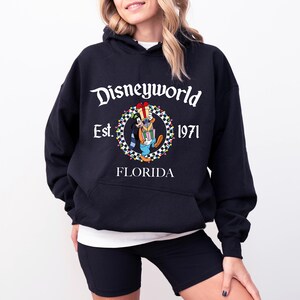 Pu&ograve; includere: Felpa con cappuccio nera con la scritta "Disneyworld" in bianco. La felpa presenta una grafica circolare con un personaggio dei cartoni animati e le parole "Est. 1971 FLORIDA". Il design &egrave; in bianco, rosso, verde, blu e giallo.