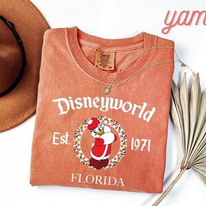 May include: A coral-coloured t-shirt featuring a Disneyworld graphic with Donald Duck in a Santa outfit. The shirt includes the text "Disneyworld Est. 1971 Florida". A gold necklace and a brown hat are also visible.