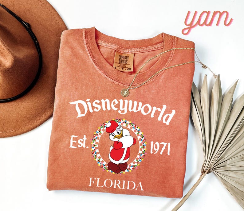 May include: A coral-coloured t-shirt featuring a Disneyworld graphic with Donald Duck in a Santa outfit. The shirt includes the text "Disneyworld Est. 1971 Florida". A gold necklace and a brown hat are also visible.