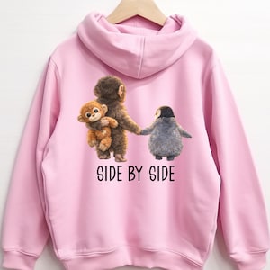 May include: Pink hooded sweatshirt featuring a graphic of three cartoon animals: a monkey holding a smaller monkey, and a penguin. The text "SIDE BY SIDE" is printed below the image in black. The sweatshirt is hanging on a wooden hanger.