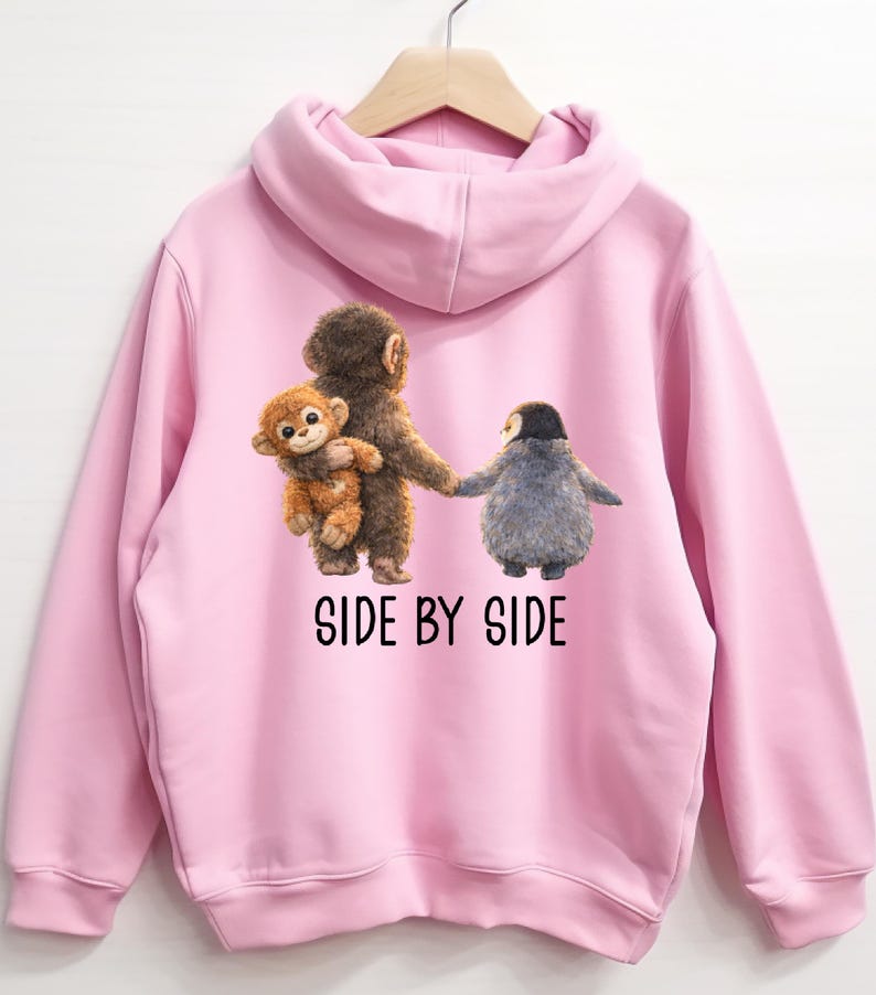 May include: Pink hooded sweatshirt featuring a graphic of three cartoon animals: a monkey holding a smaller monkey, and a penguin. The text "SIDE BY SIDE" is printed below the image in black. The sweatshirt is hanging on a wooden hanger.