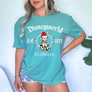 May include: Teal t-shirt with a Disneyworld graphic. The design includes the text "Disneyworld Est. 1971 FLORIDA" and a cartoon character in a Santa hat. The shirt is short-sleeved and casual, suitable for everyday wear.