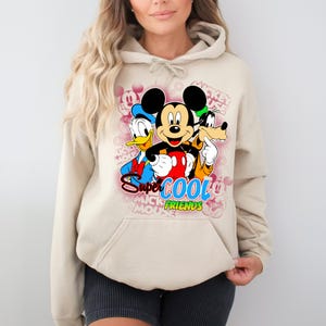 Puede incluir: Sudadera con capucha beige con un gr&aacute;fico colorido de Mickey Mouse, el Pato Donald y Goofy. El gr&aacute;fico incluye el texto "Super Cool Friends". La sudadera tiene un bolsillo delantero y mangas largas.