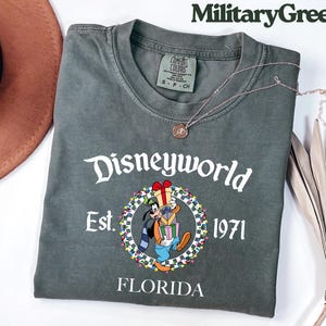 May include: Military green t-shirt with "Disneyworld" in white script. The shirt features a graphic of Goofy holding gifts, surrounded by a wreath, with "Est. 1971" and "Florida" below. A silver necklace with a pendant is draped on the shirt.