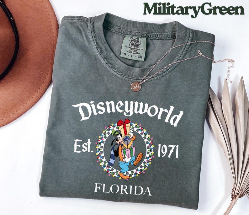 May include: Military green t-shirt with "Disneyworld" in white script. The shirt features a graphic of Goofy holding gifts, surrounded by a wreath, with "Est. 1971" and "Florida" below. A silver necklace with a pendant is draped on the shirt.