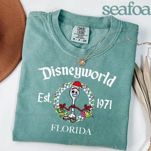 May include: Seafoam green t-shirt with the text "Disneyworld Est. 1971 Florida". The shirt features a cartoon fork character wearing a Santa hat and holding Christmas ornaments. A gold necklace is draped across the shirt.